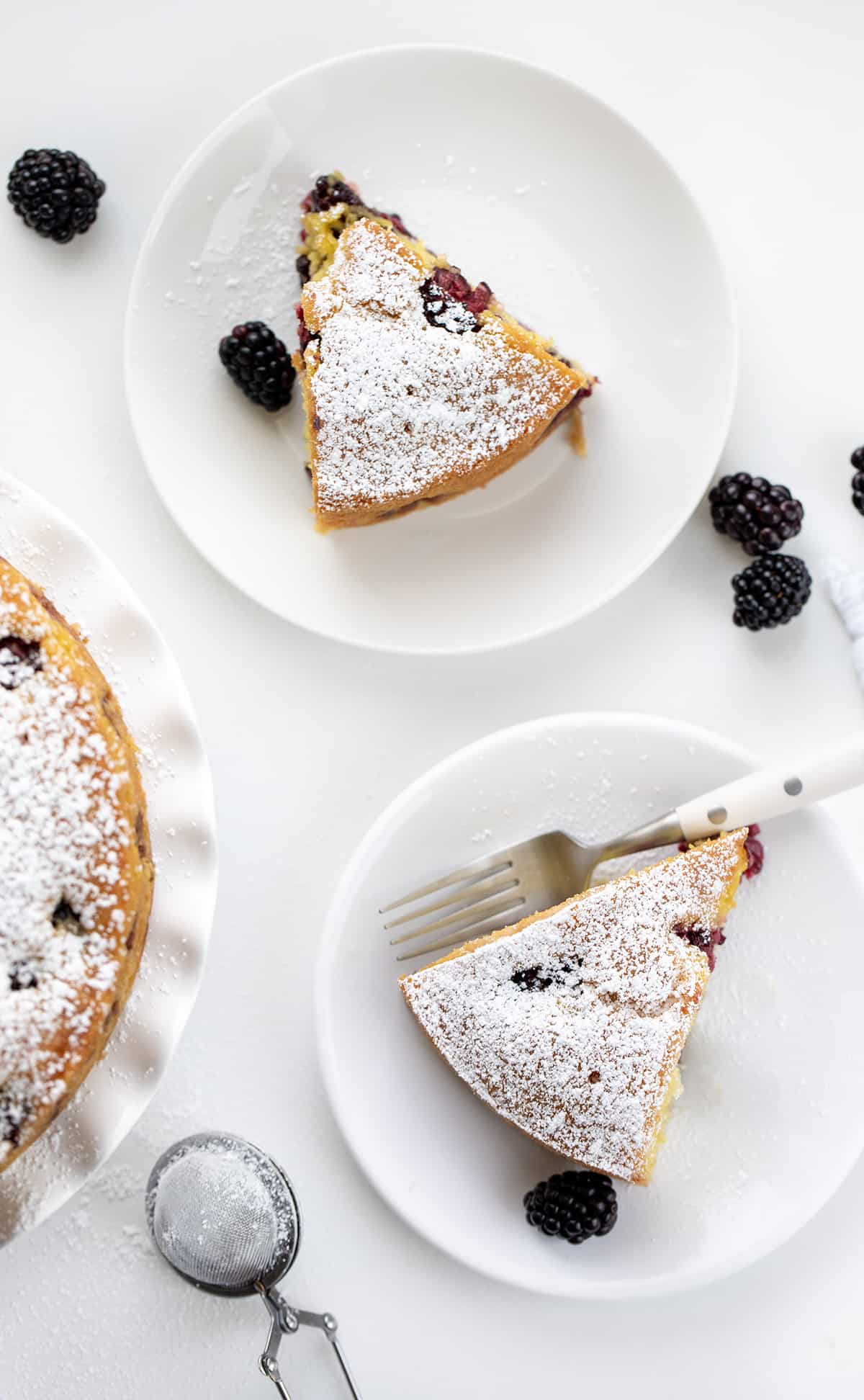 White Plates with Blackberry Breakfast Cake on Them with White Form and Fresh Blackberries on a White Counter.