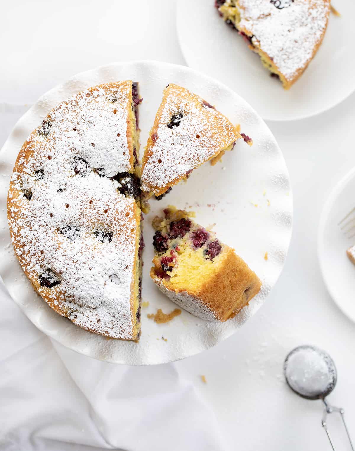 Blackberry Breakfast Cake on a Cake Stand with Some Pieces Removed and One Piece on its Side and Other plates Around. 