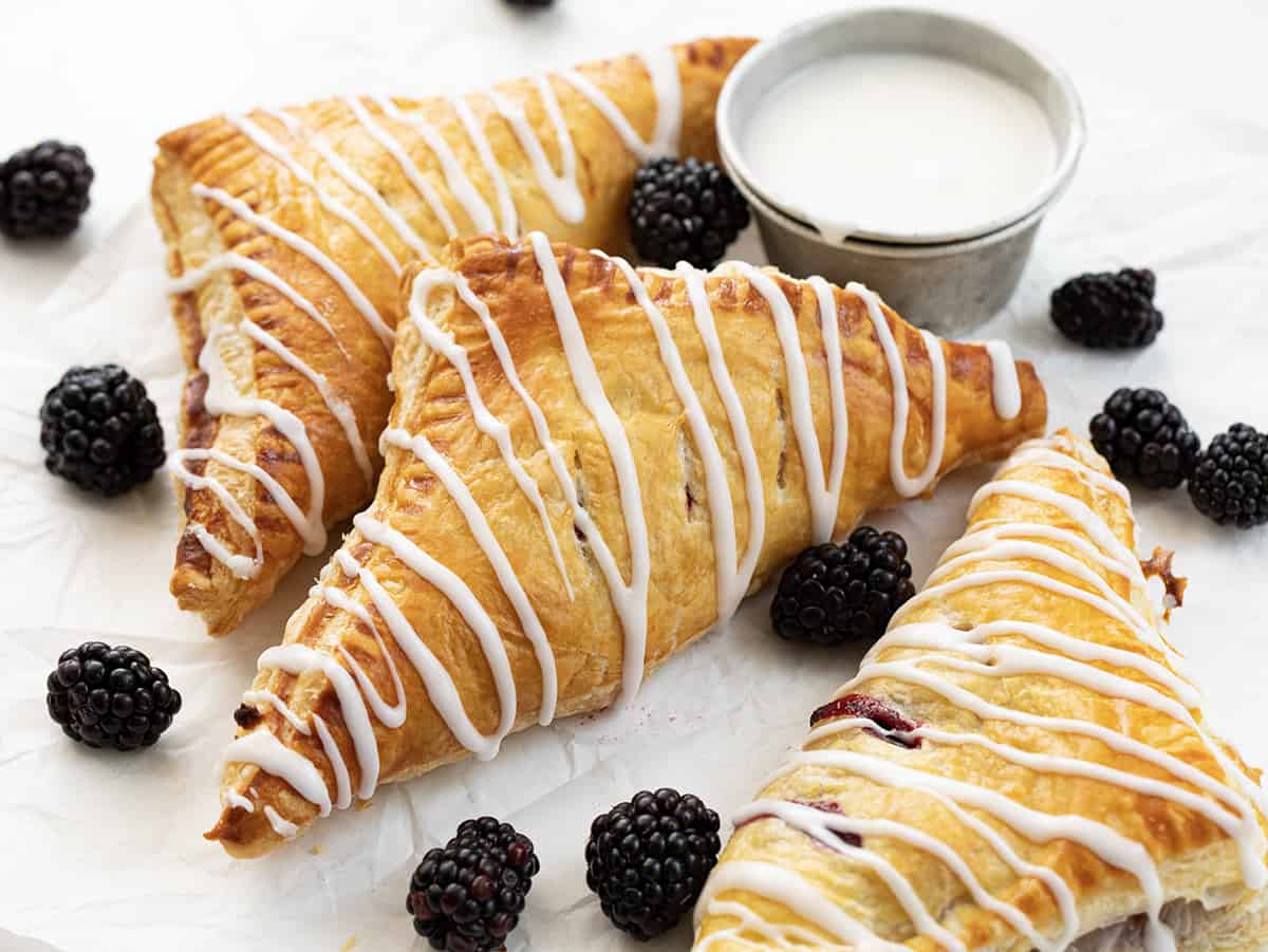 Blackberry Cream Cheese Turnovers on a Pan with Glaze and a Few Blackberries on a White Counter.