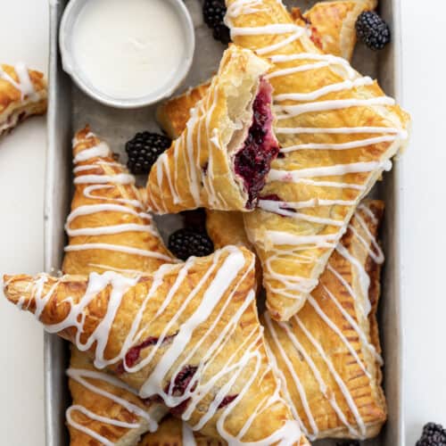 Blackberry Cream Cheese Turnovers on a Pan with Glaze and a Few Blackberries on a White Counter.