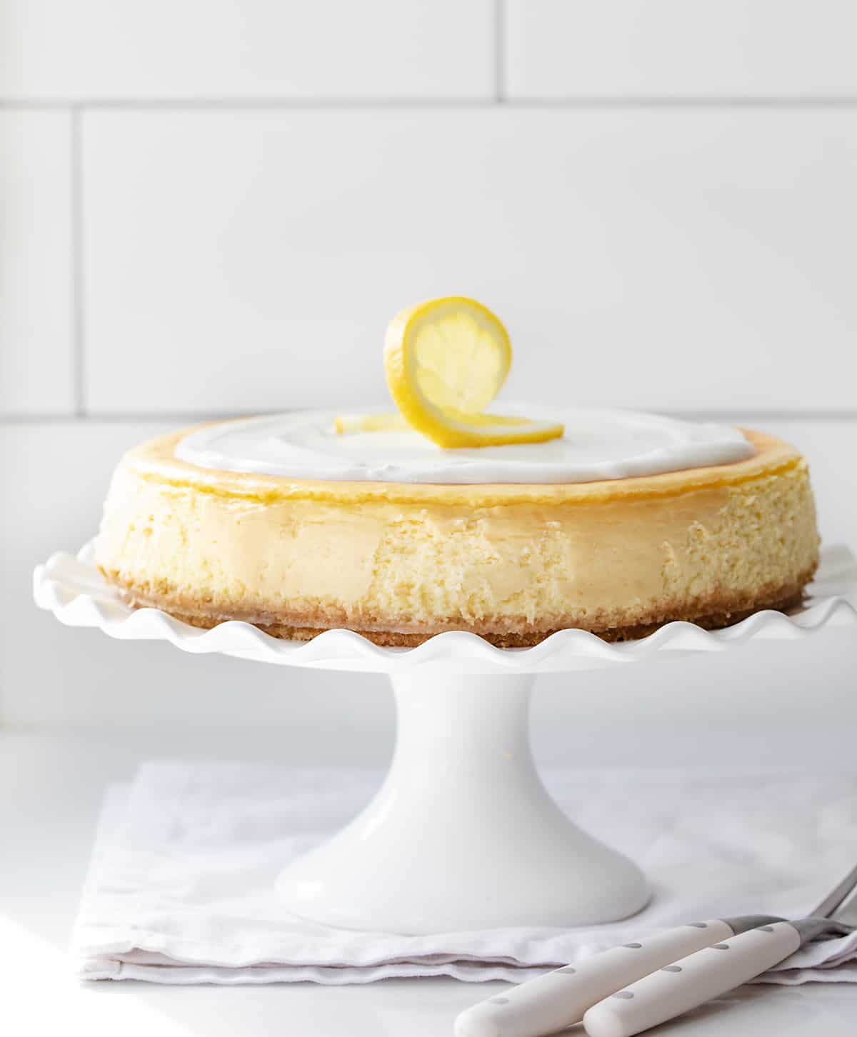 Lemon Cheesecake on a White Cake Stand with a Ruffled Edge on a White Counter in Front of a Subway Tiled Wall. 