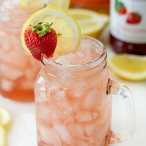 Glasses of Whiskey Strawberry Lemonade on a White Counter with Strawberry Syrup in the Background.