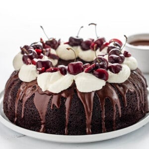 Whole Black Forest Bundt Cake on a White Serving Platter on a White Counter with Chocolate Sauce in Background.