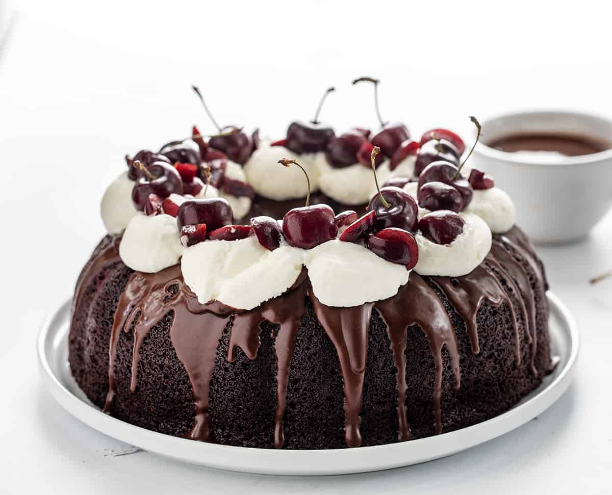 Whole Black Forest Bundt Cake on a White Serving Platter on a White Counter with Chocolate Sauce in Background.
