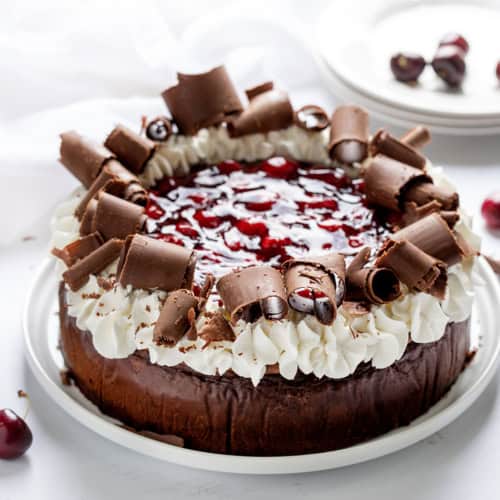 Black Forest Cheesecake on a White Cake Plate on a White Surface and White Plates in the Background.