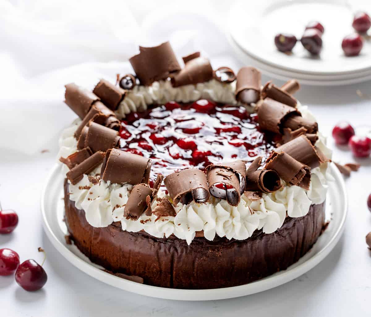 Black Forest Cheesecake on a White Cake Plate on a White Surface and White Plates in the Background.