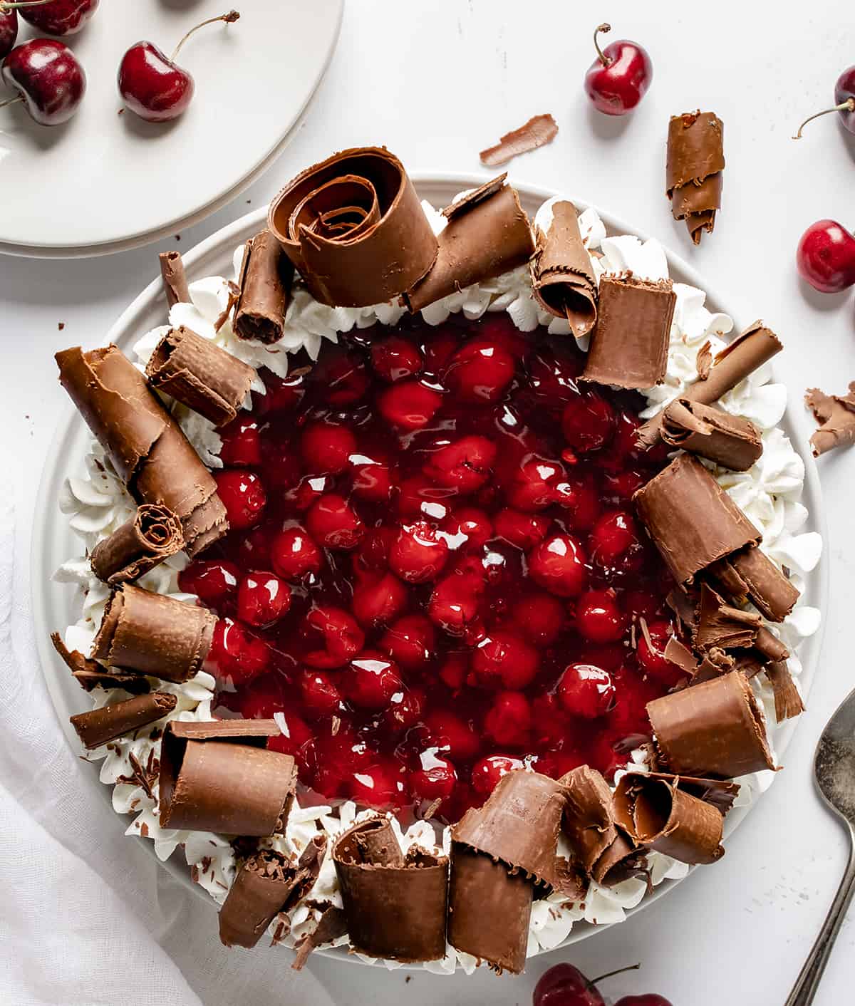 Black Forest Cheesecake on a White Surface from Overhead with Some White Plates and Cherries Around.