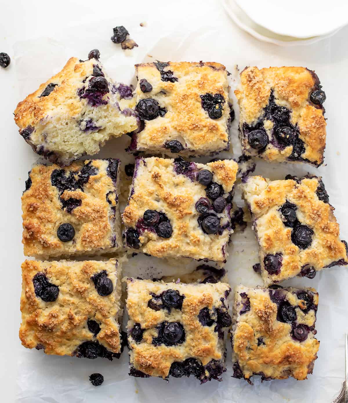 Blueberry Butter Swim Biscuits combine the flavors of blueberries, butter, and a hint of sweetness with a moist and fluffy crumb and crispy golden brown top. They can be enjoyed as a breakfast treat, a snack, or even as a dessert! Plus, they are SO EASY to make! Blueberry Butter Swim Biscuits on a White Counter from Overhead and Cut Into Pieces.