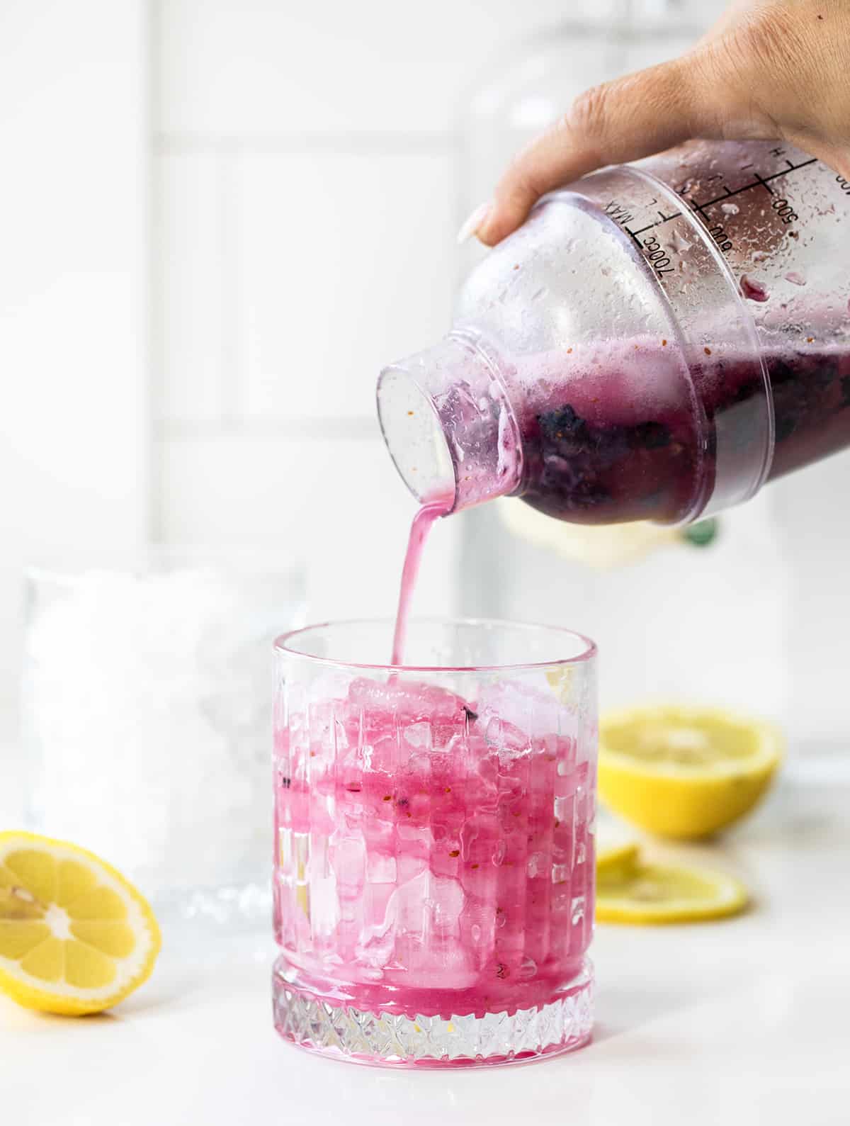 Pouring Freshly Shaken Spiked Blueberry Lemonade from Shaker Into Glass with Ice in It on a White Counter with Lemons.
