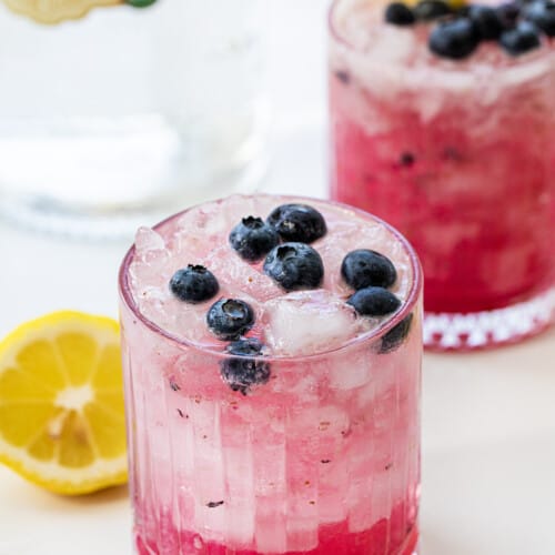Glasses of Spiked Blueberry Lemonade in Front of a Vodka Bottle with Fresh Lemon and Blueberries.
