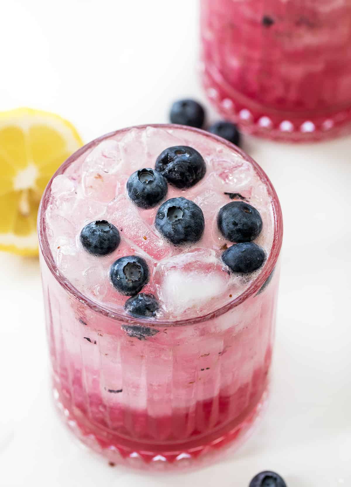 Top of a Glass of Spiked Blueberry Lemonade Surrounded by Another Glass, Lemons, and Fresh Blueberries on a White Counter.