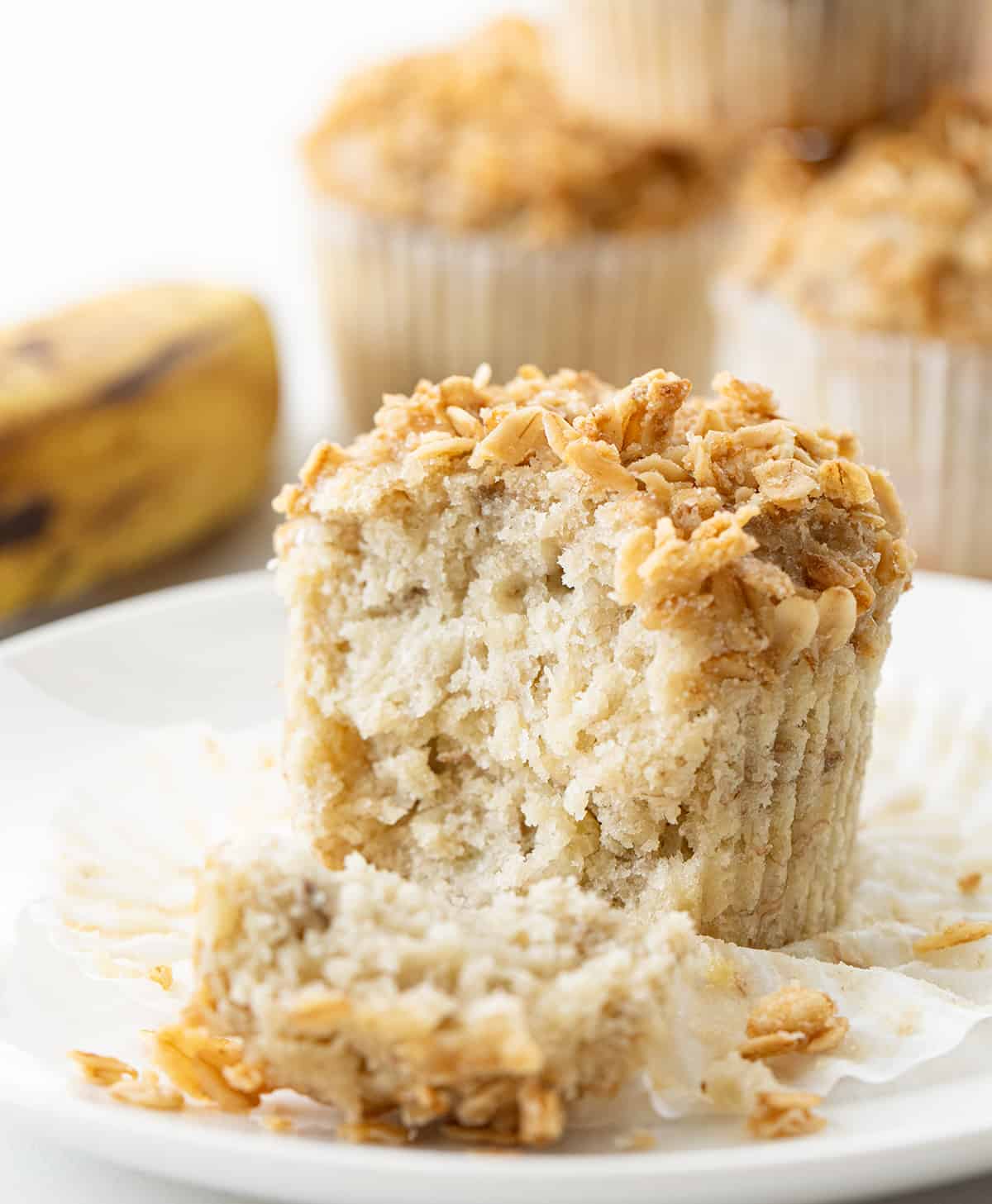 One Banana Crunch Muffin on a Counter with Some Stacked Behind Near a Banana and Some of the Muffin Removed Showing Inside Texture.
