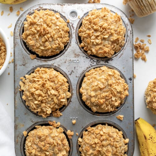 Banana Crunch Muffins in a Muffin Pan with Bananas and More Muffins Around Looking Down from Overhead.