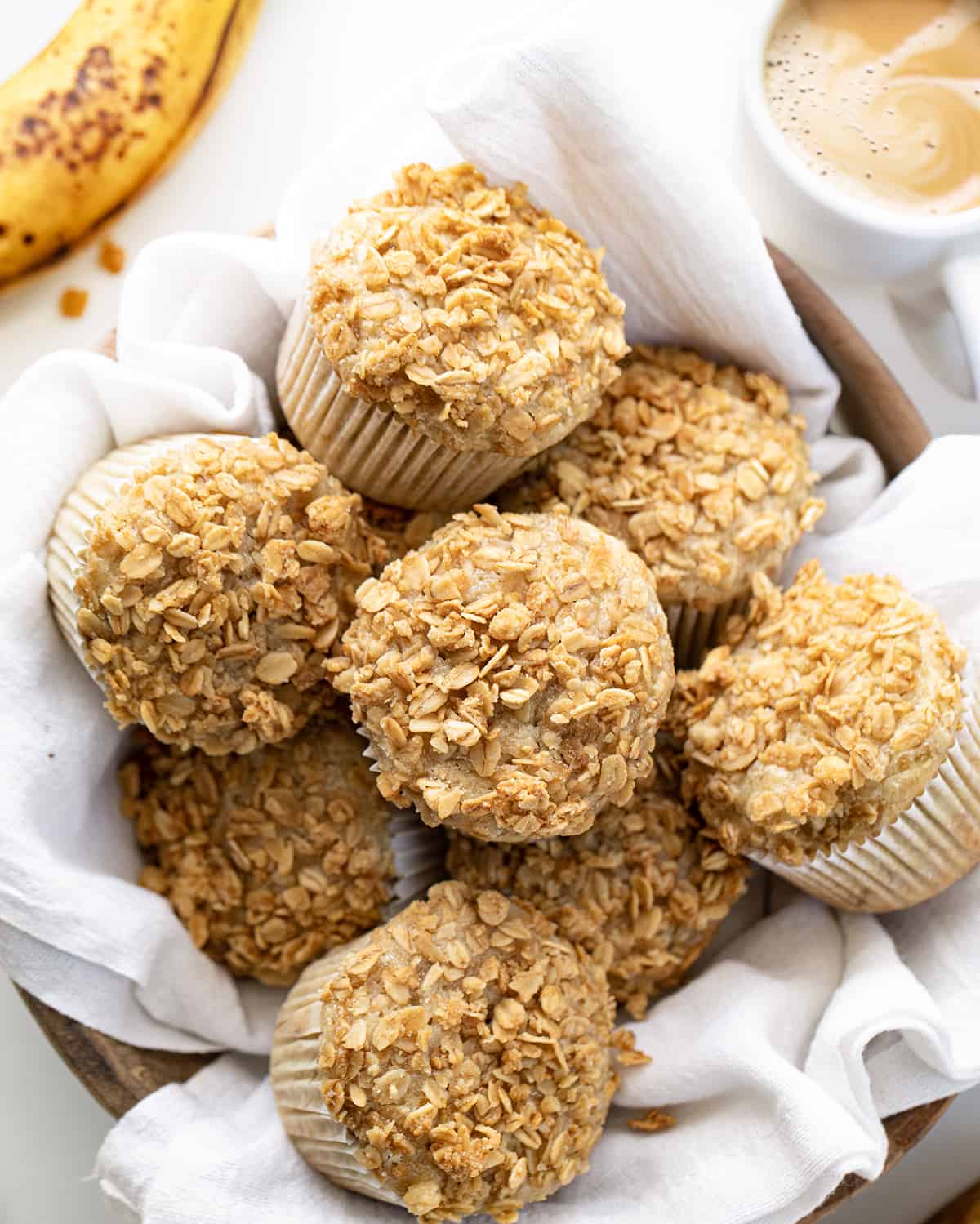 Banana Crunch Muffins in a Bowl on a White Towel with Banana and Coffee Around it on a White Table. 