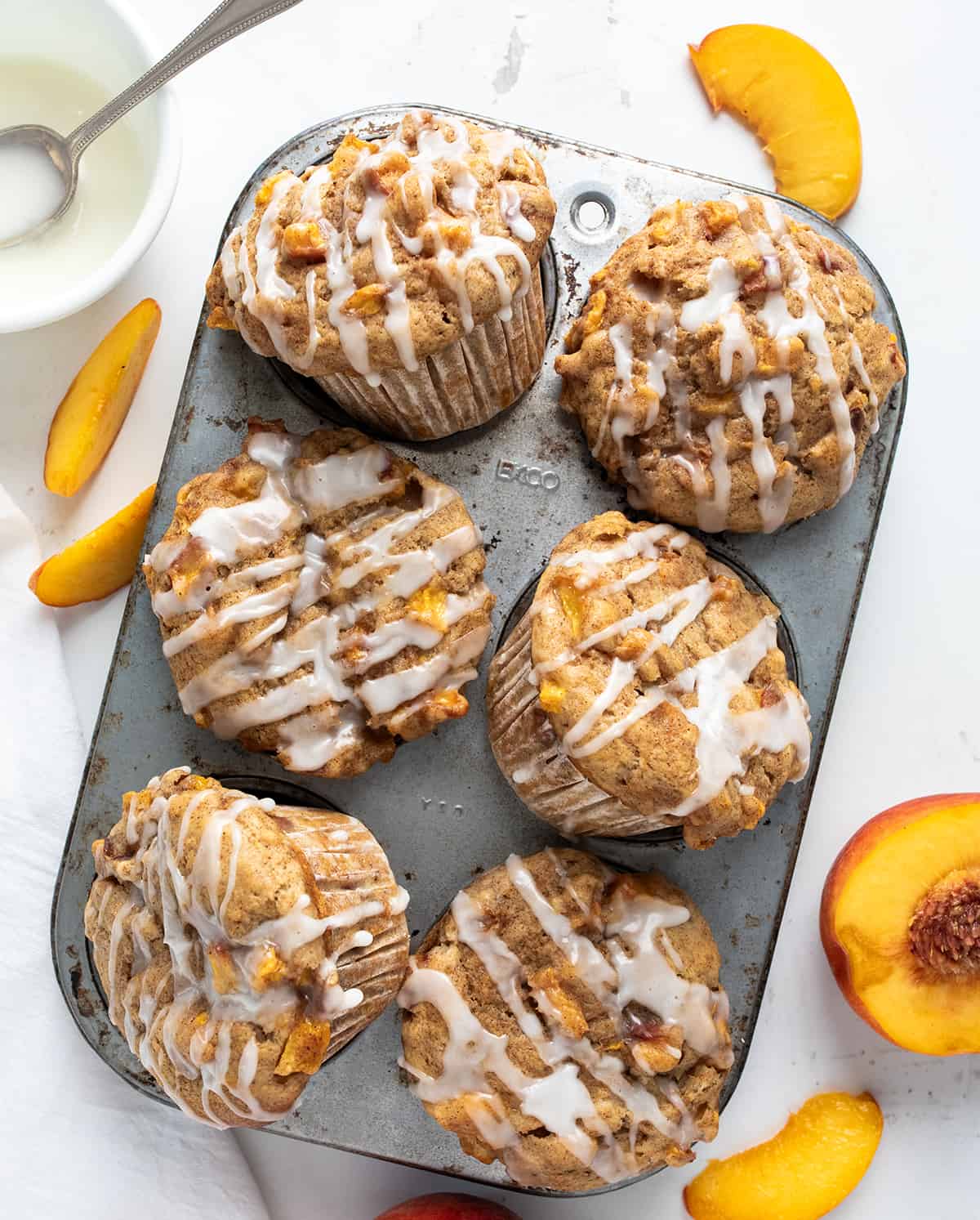 Peach Muffins in a Muffin Tin Next to Peaches on a White Counter.