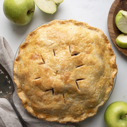 Whole Bourbon Apple Pie on a White Table with Apples and Towel.