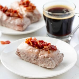 Maple Bacon Donut Stick on a White Plate with Coffee and More Donuts in the Background.