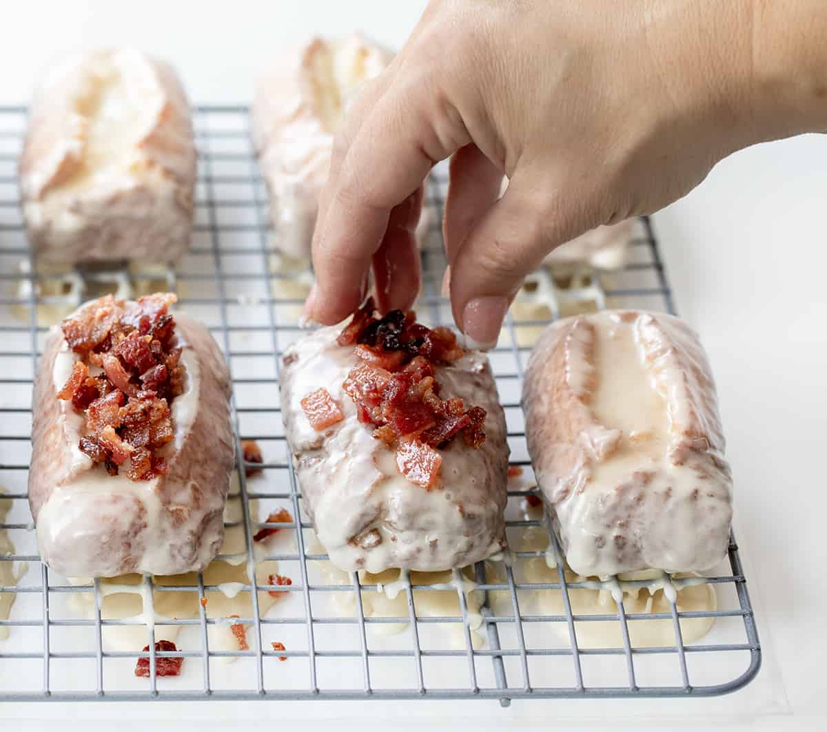 Hand Dropping Bacon Bits onto Freshly Glazed Donut Sticks with a Maple Glaze.