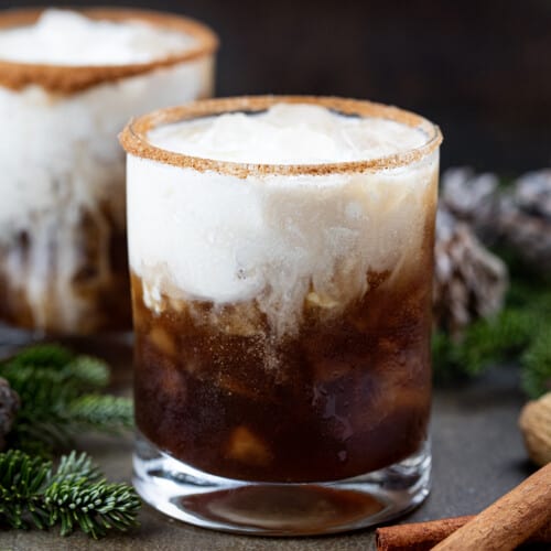Two Gingerbread White Russian cocktails on a dark table with cinnamon sticks and evergreens around.