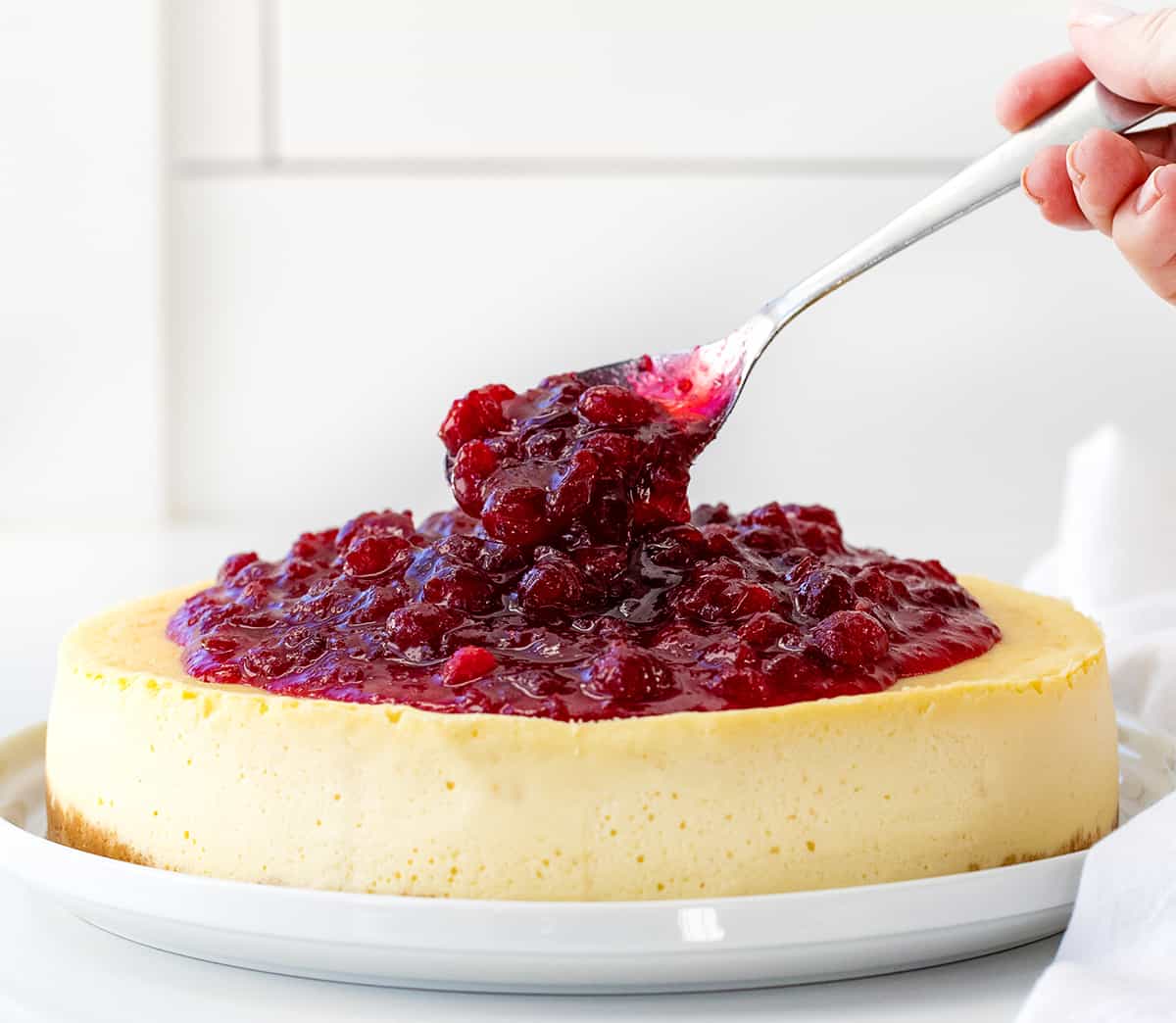 Spooning cranberry sauce over plain cheesecake on a white counter.