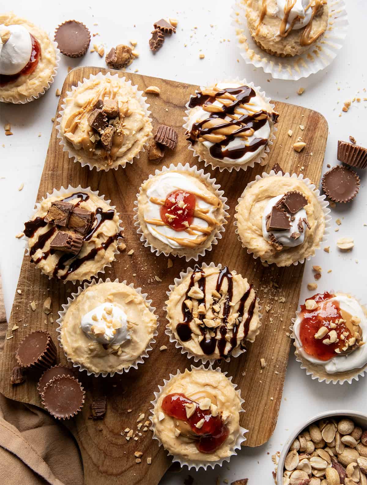 Variety of decorated Mini Peanut Butter Cheesecakes on a cutting board on a white counter with reeses, jelly, whipped cream, and peanuts.