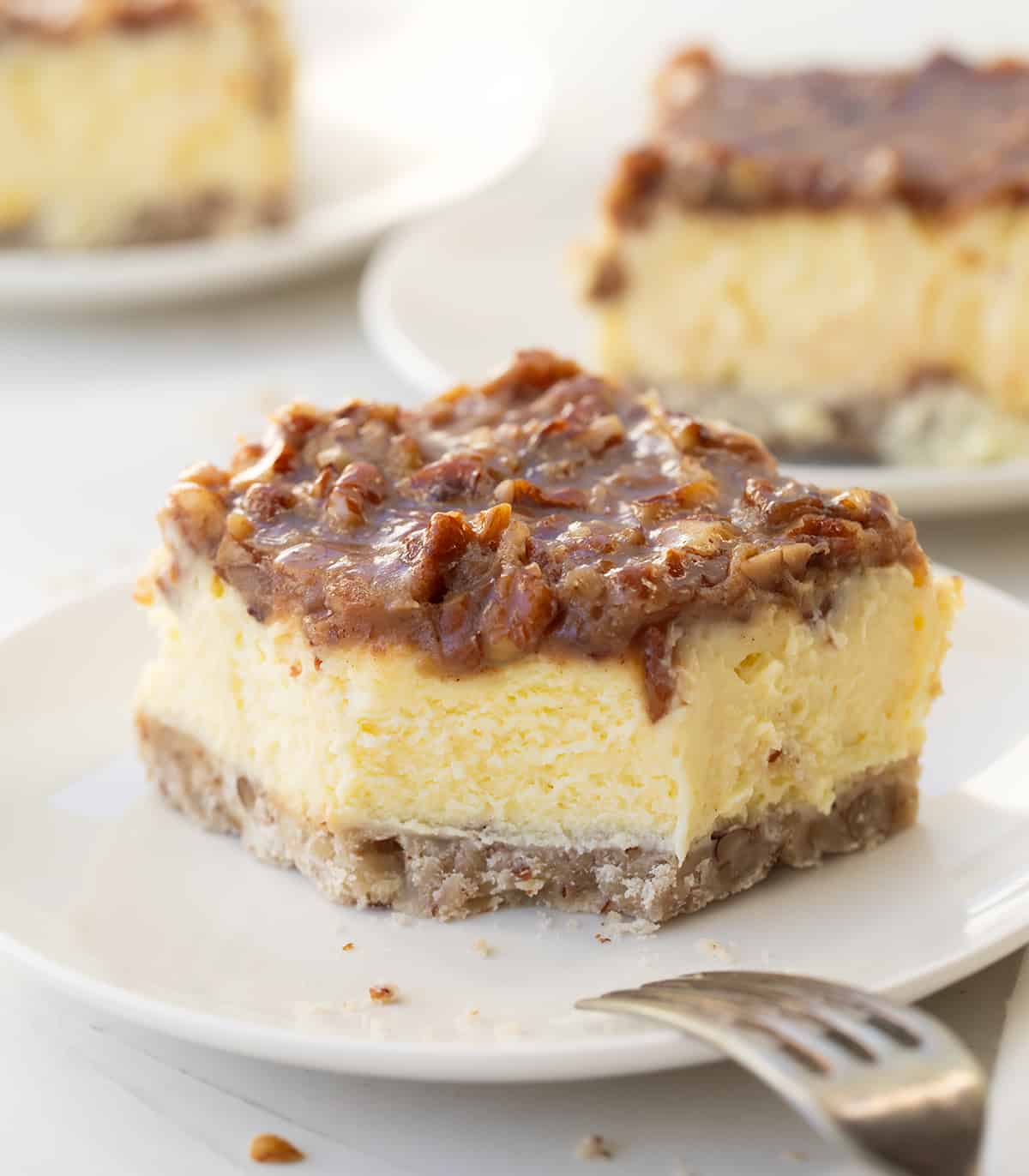 One Pecan Pie Cheesecake Bar on a plate with a bite removed and fork resting on the white plate.