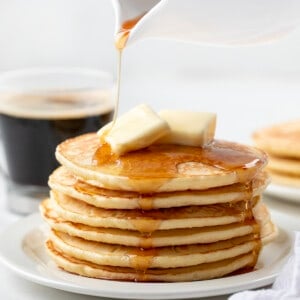 Pouring syrup over a stack of Homemade Bisquick Pancakes on a white plate.