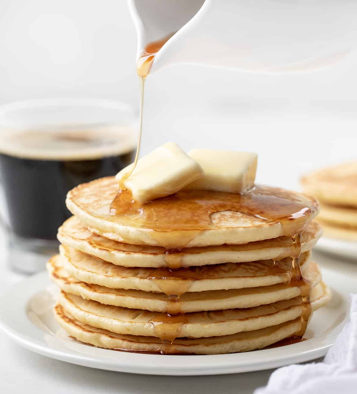 Pouring syrup over a stack of Homemade Bisquick Pancakes on a white plate. 
