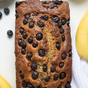 Loaf of Blueberry Banana Bread baked and on a white counter with blueberries and bananas on the sides from overhead.