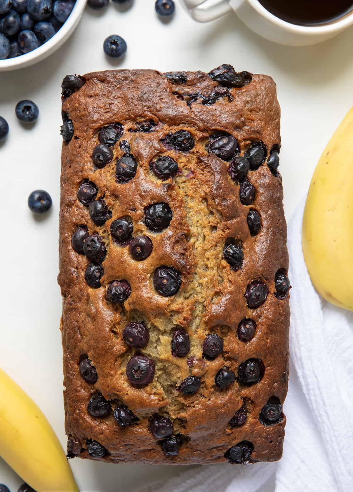 Loaf of Blueberry Banana Bread baked and on a white counter with blueberries and bananas on the sides from overhead.