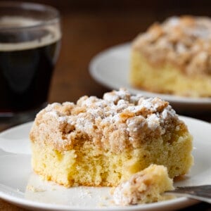 Piece of Crumb Cake on a plate with a bite removed and coffee in the background.