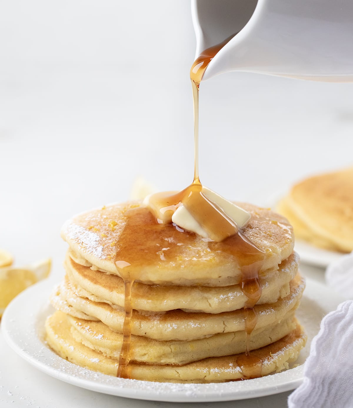 Pouring syrup over a stack of Lemon Ricotta Pancakes on a white plate.