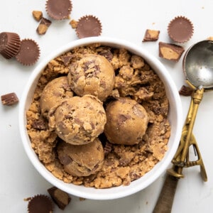Bowl of Edible Peanut Butter Cookie Dough on a white table with some scoops in the bowl and an ice cream scoop next to it.