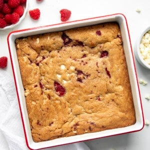Pan of Raspberry White Chocolate Blondies on a white counter with raspberries and white chocolate morsels from overhead.