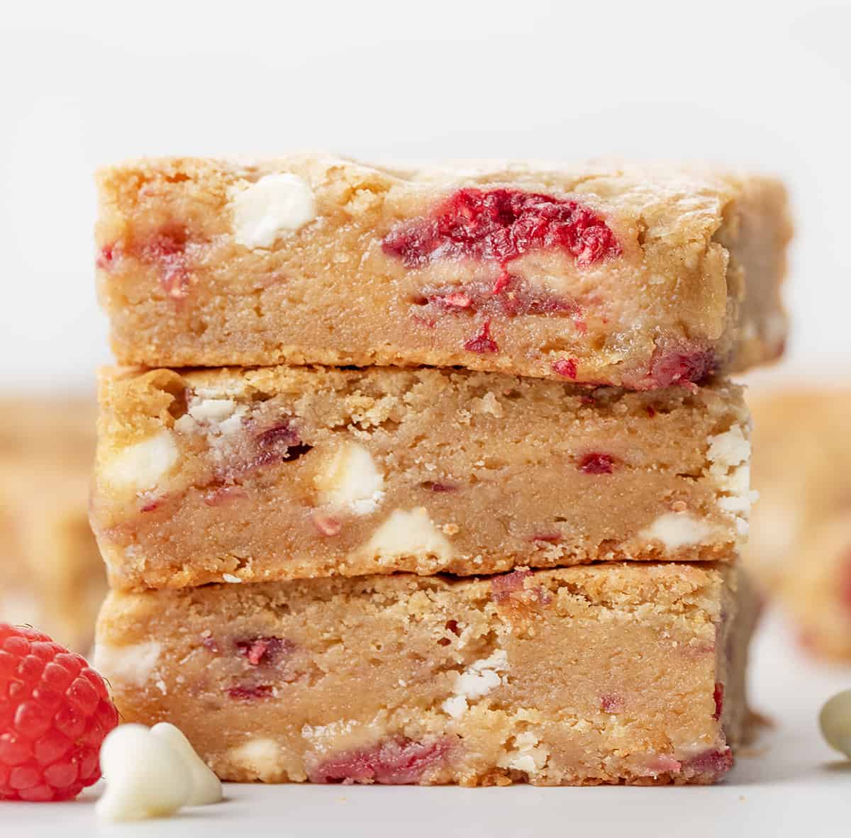 Stack of Raspberry White Chocolate Blondies on a white counter with a raspberry and white chocolate morsels.