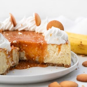 Banana Pudding Cheesecake on a cake plate with some pieces removed showing the caramel dripping down the sides.