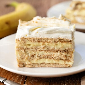 One piece of Banana Icebox Cake on a white plate near a banana and another plate.