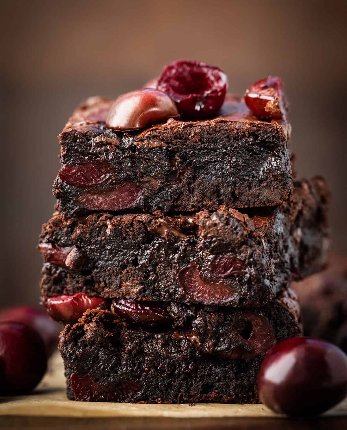 Stack of three Roasted Cherry Brownies on a wooden table surrounded by fresh cherries.