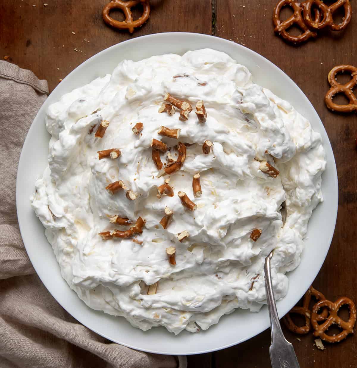 White bowl filled with Pineapple Pretzel Fluff with broken pretzels on top and surrounded by whole pretzels.