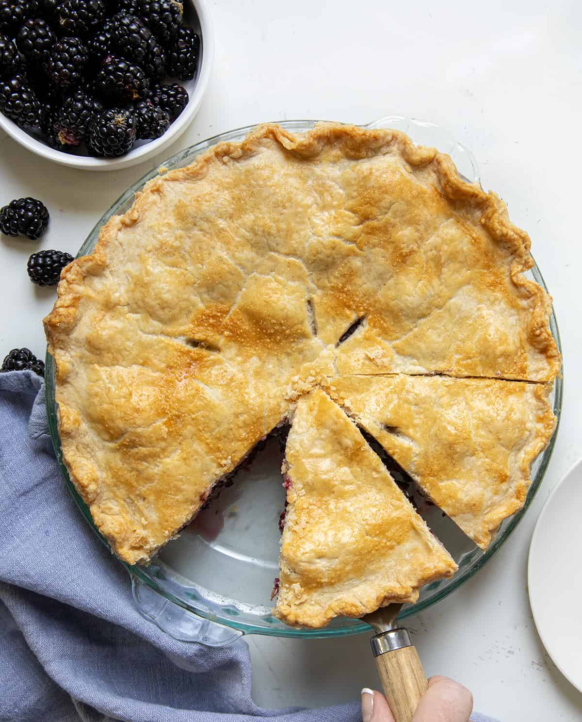 Whole Blackberry Pie on a white counter with one piece missing and another piece being picked up from overhead.