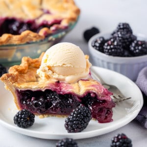 Piece of blackberry pie on a white plate with ice cream on top.
