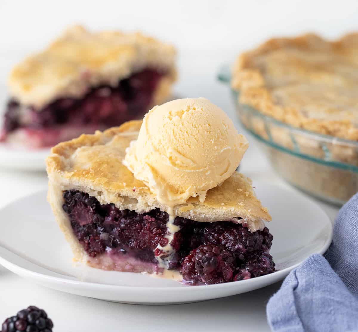 Piece of Blackberry Pie on a white plate in front of another piece and the whole pie on a white counter.