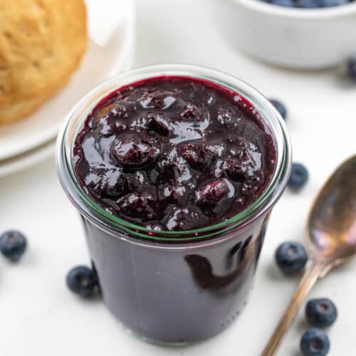 Jar of Blueberry Jam next to a spoon with a biscuit and fresh blueberries in the background.