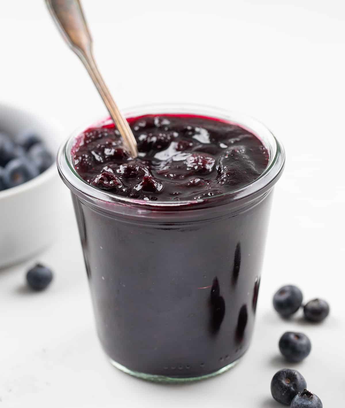 Jar of Blueberry Jam with a spoon in it on a white table. 