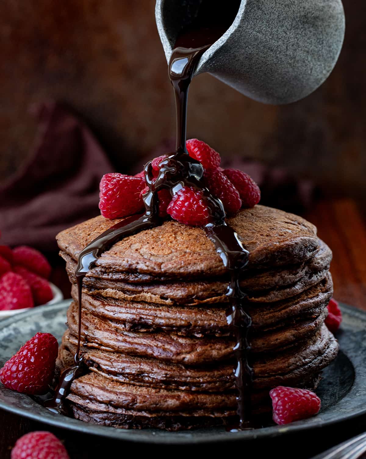 Pouring chocolate sauce over a stack of Chocolate Pancakes on a wooden table with raspberries on top and around.