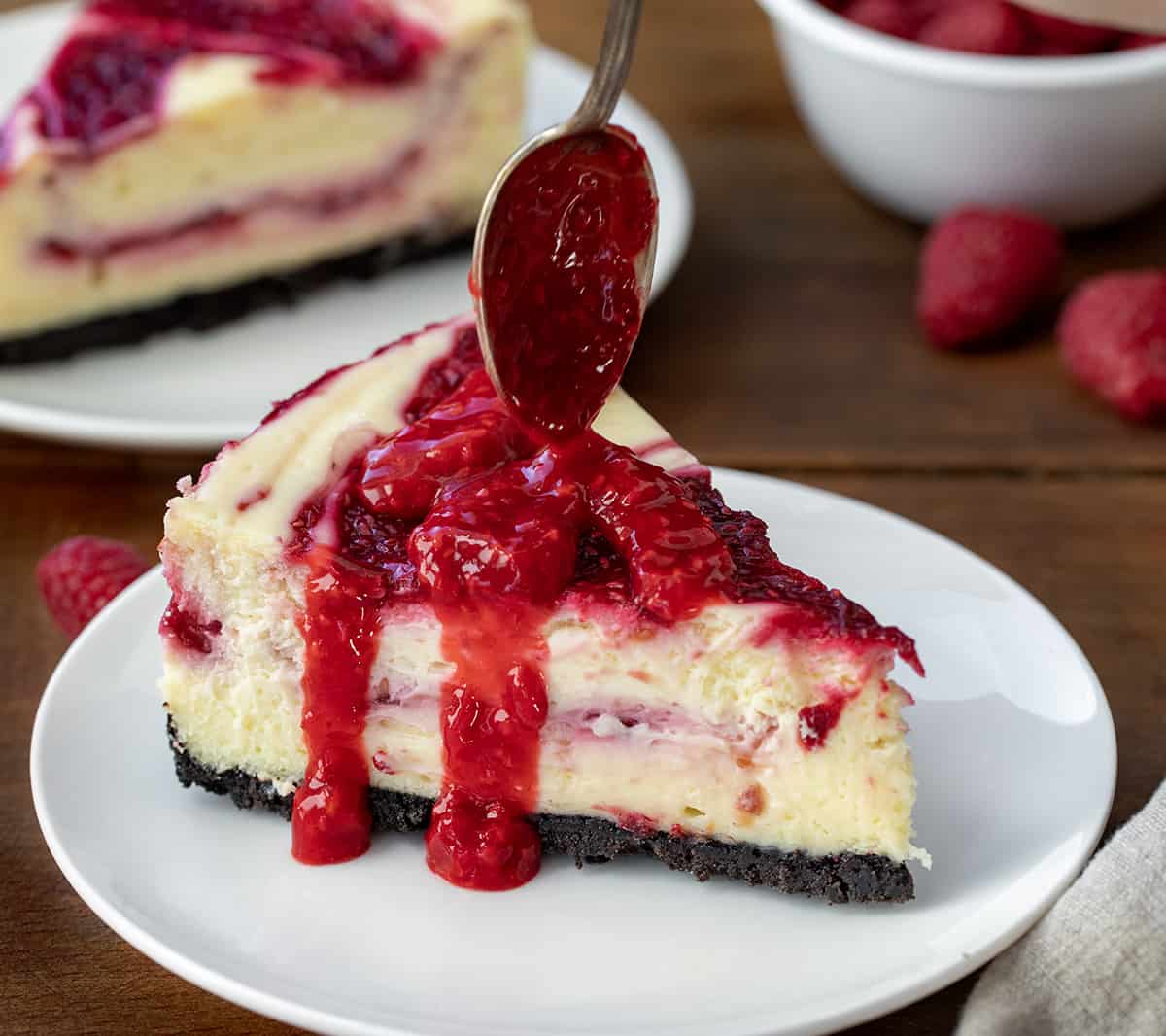 Piece of White Chocolate Raspberry Cheesecake on a white plate with raspberry sauce being drizzled over top on a wooden table.