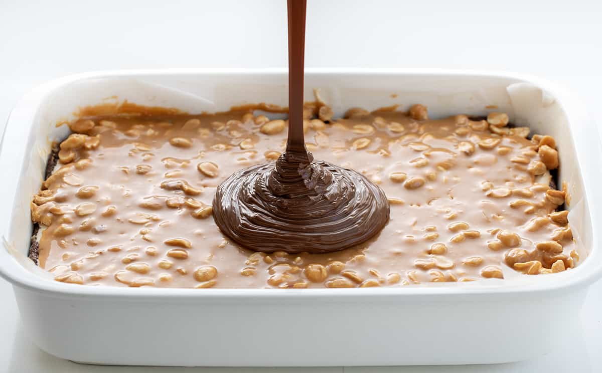 Pouring chocolate onto peanut butter caramel in a pan of Snickers Brownies.