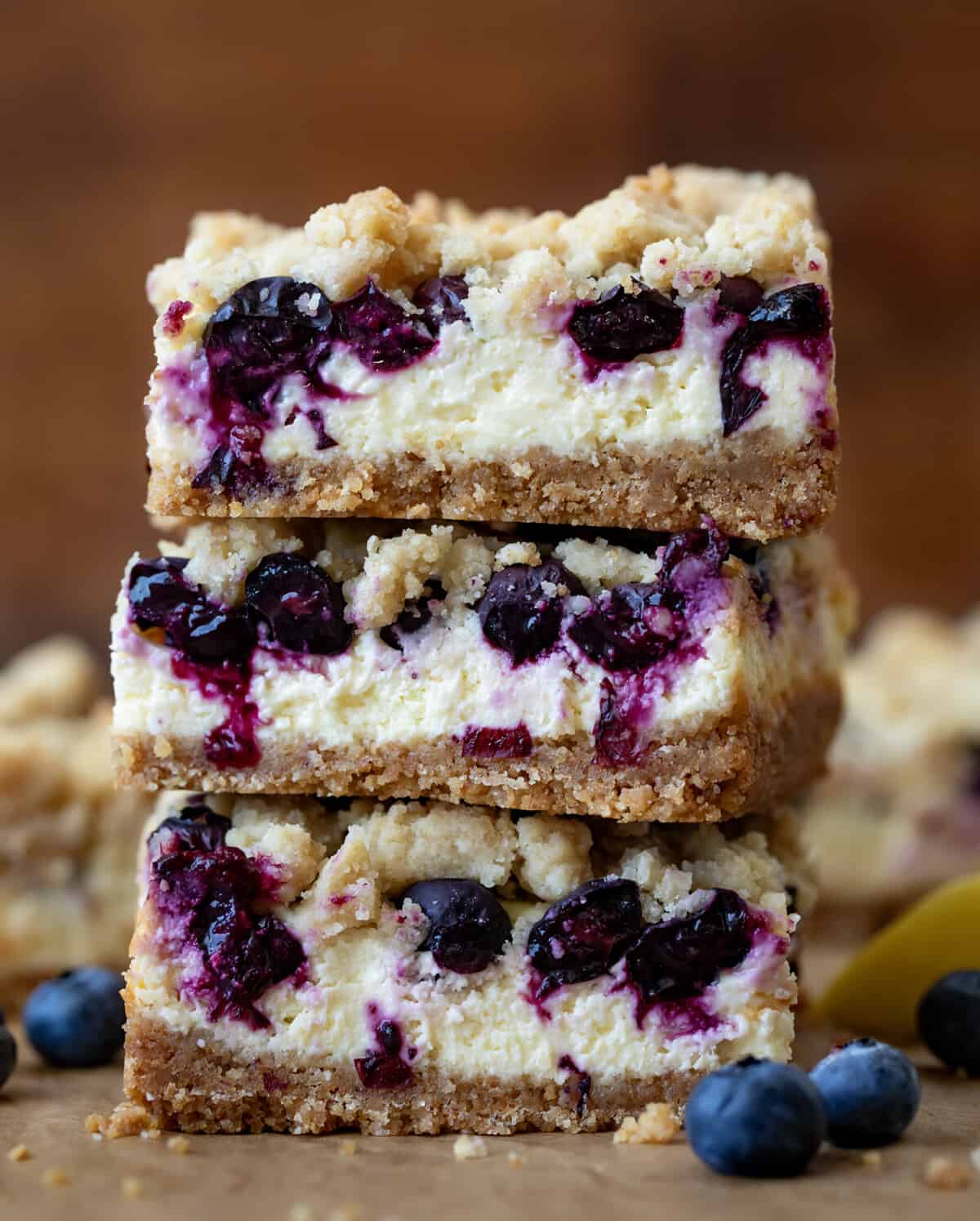 Blueberry Lemon Cheesecake Bars are a graham cracker crust topped with a creamy lemon cheesecake filling, fresh blueberries, and a crumble topping. They are baked until the topping is golden brown. Then, the bars are chilled until set before being cut and served. There is no wrong time to enjoy one of these bars, from a treat with a cup of coffee to a dessert that everyone will love! Stack of Blueberry Lemon Cheesecake Bars on a wooden table.