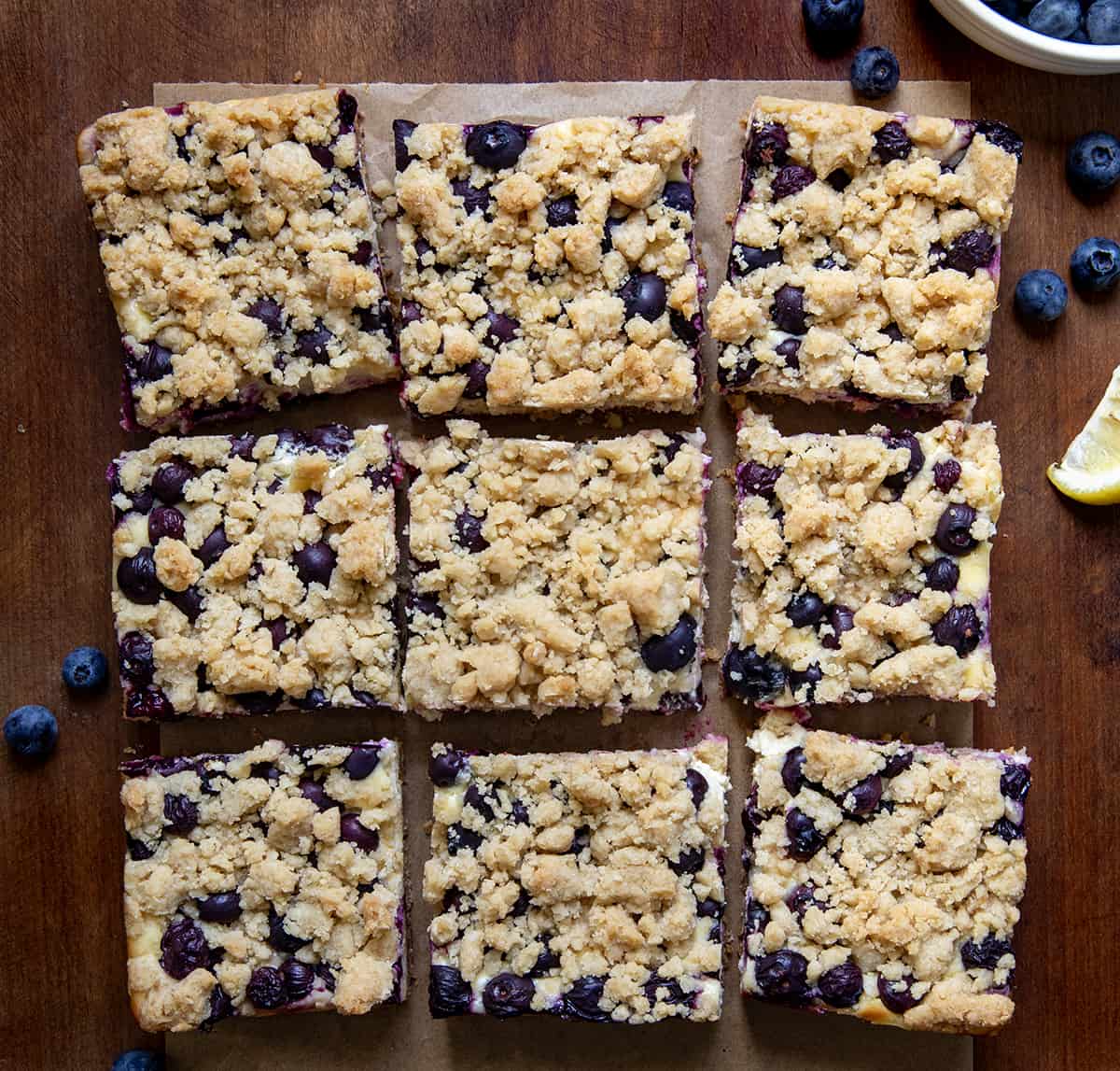 Blueberry Lemon Cheesecake Bars on a wooden table cut into pieces with blueberries and fresh lemon.