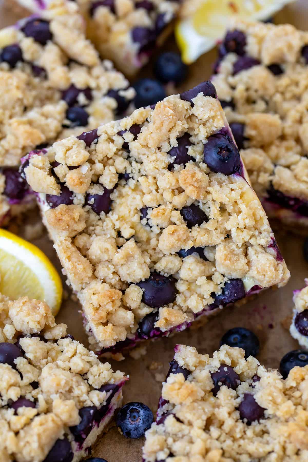 Blueberry Lemon Cheesecake Bars are a graham cracker crust topped with a creamy lemon cheesecake filling, fresh blueberries, and a crumble topping. They are baked until the topping is golden brown. Then, the bars are chilled until set before being cut and served. There is no wrong time to enjoy one of these bars, from a treat with a cup of coffee to a dessert that everyone will love! Close up of Blueberry Lemon Cheesecake Bars all stacked together on a wooden table.